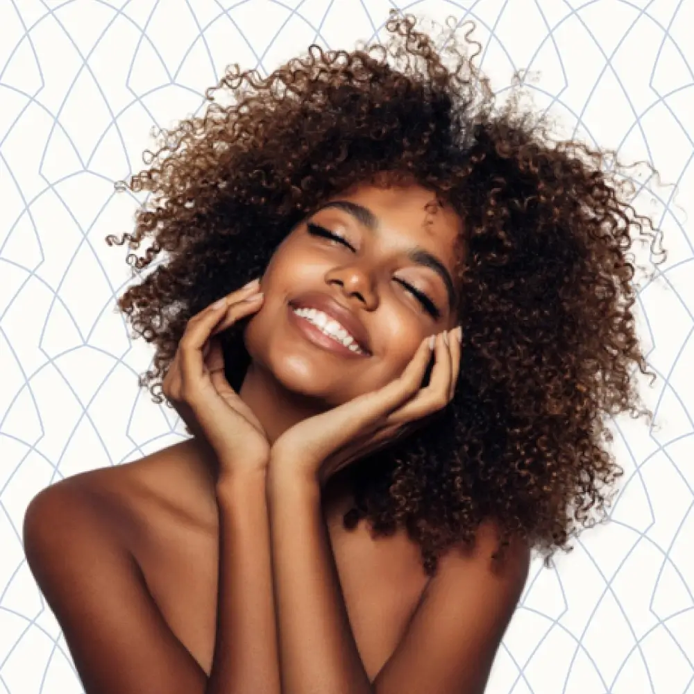 Young woman with curly hair smiling and posing with both hands on her face | Hair Restoration | Andromeda Medspa